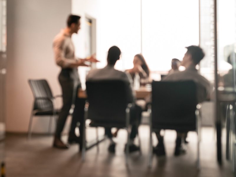 Business Presentation, Blurred Background. Businessman Giving Speech During Seminar With Coworkers In Office, Standing At Desk In Boardroom, Diverse People Sitting At Table And Listening To Speaker